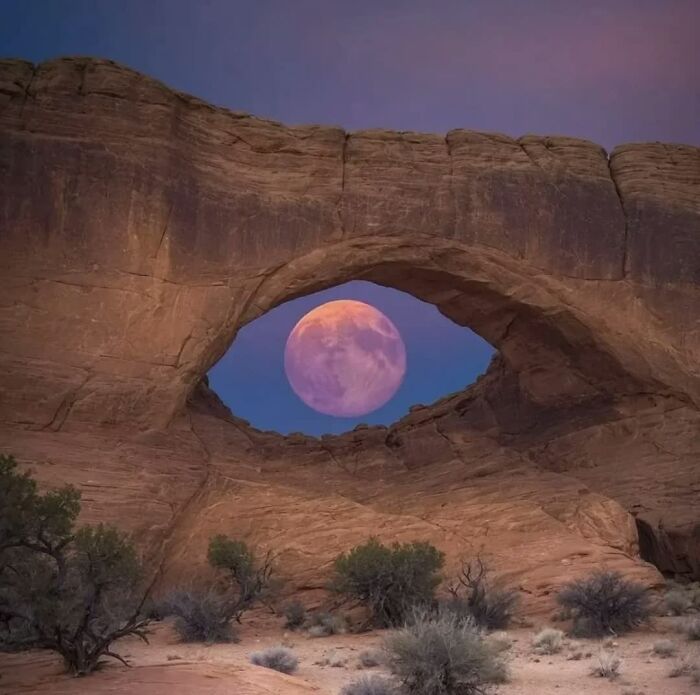 A stunning red moon seen through a natural rock arch, a weird image that lives rent-free in your mind.