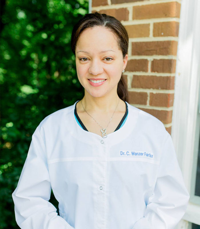 Young woman wearing a white lab coat with a necklace, standing outdoors near a brick wall, related to Justin Fairfax case.