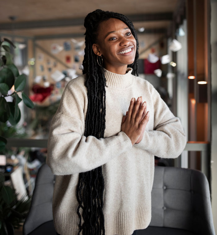 A young Black woman with long dreadlocks, wearing a cozy sweater, smiling with hands pressed together. Favorite things about Black people.