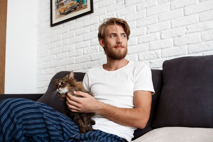 Young man sitting on couch holding a cat, reflecting a first date with red flags and instant regret vibes.