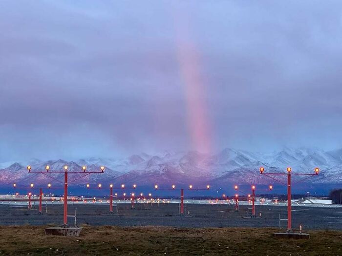 An airport runway with red lights, snowy mountains, and a pink light beam in the sky, illustrating life in Alaska.