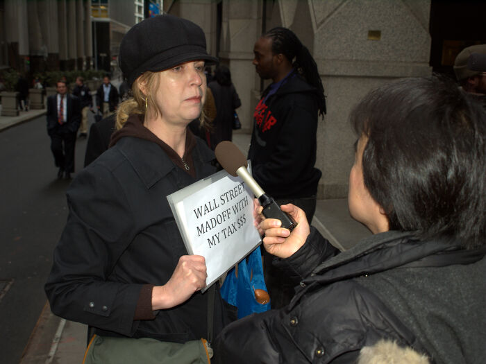 Woman holding sign about Wall Street and Madoff during a street interview, representing events from the year you were born.