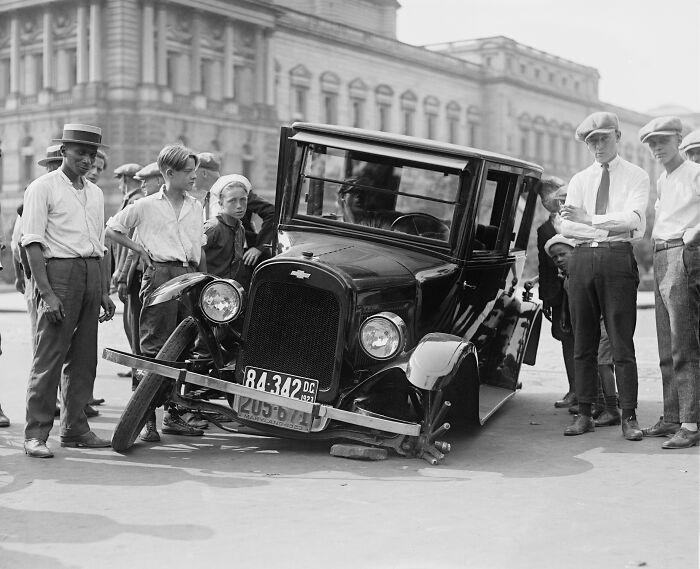 Group of people gathered around a vintage car with a flat tire in a rare and interesting photo from a past time period.