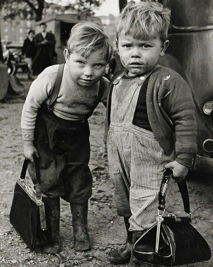 Two young boys, one in suspenders and the other in overalls, holding bags, offering a fascinating photo of history.