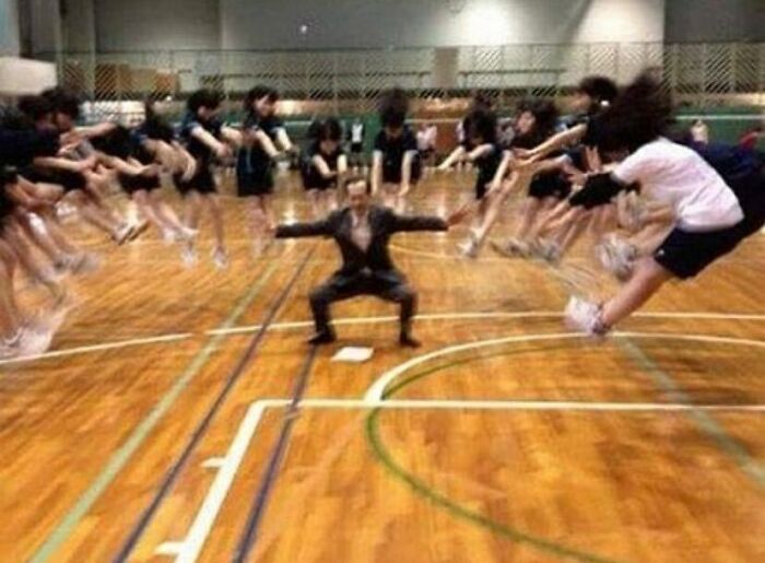 A cursed image of a man in a suit squatting with arms out, surrounded by students in mid-jump creating a blur effect.