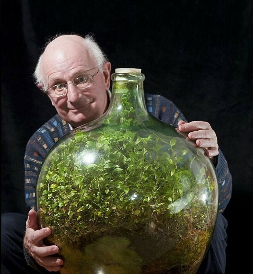 A smiling man in glasses holds a large glass bottle with a vibrant ecosystem of plants inside. Fascinating photos of history.