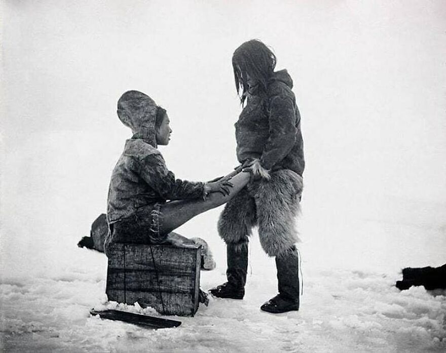 Fascinating photo showing two people in historical winter clothing on snow, one sitting on a crate, offering a glimpse into history.