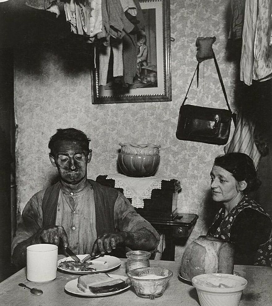 Black and white photo shows a coal miner, face smudged, eating dinner as a woman watches. Fascinating photos of history.