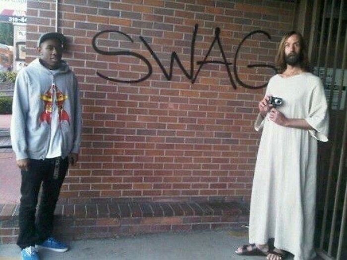A cursed image of a man in a hoodie and a man dressed as Jesus with a camera, in front of a brick wall with "SWAG" graffiti.