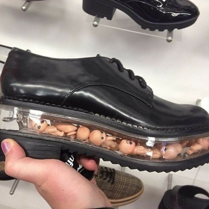 A hand holding a black dress shoe with tiny doll heads embedded in its clear sole, a truly cursed image.