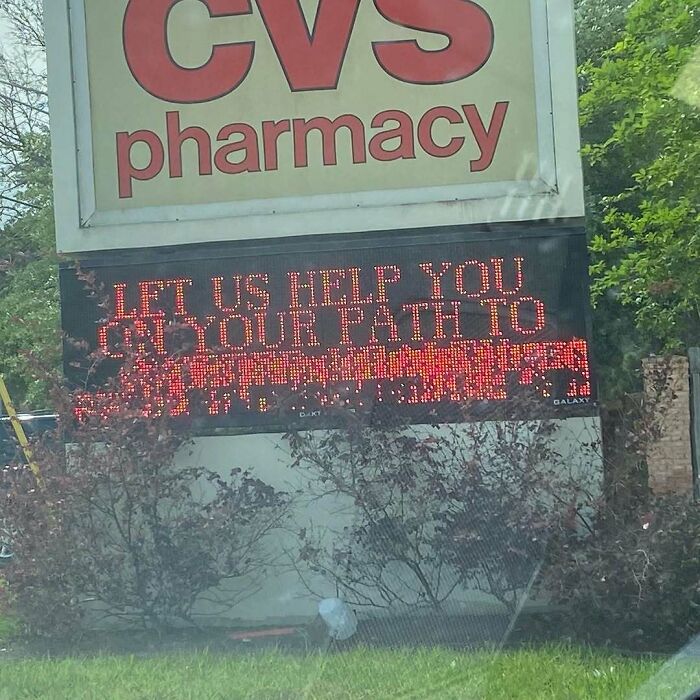 A CVS Pharmacy sign with a digital display showing garbled, cursed images text beneath a clear "LET US HELP YOU ON YOUR PATH TO".