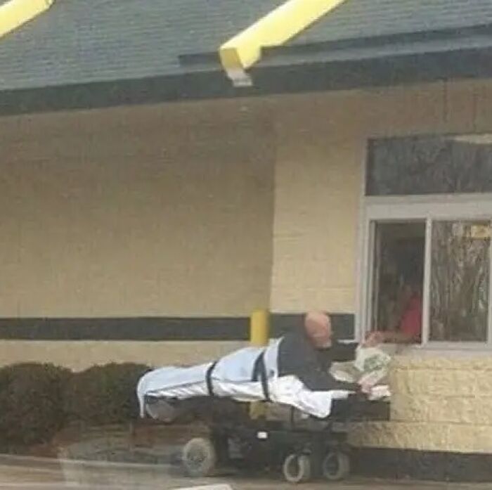 A bald man on a stretcher at a drive-thru window, receiving food. A cursed image that confuses and amuses.