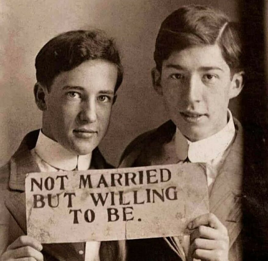Two young men in vintage attire holding a sign, "NOT MARRIED BUT WILLING TO BE." A fascinating photo that teaches about history.