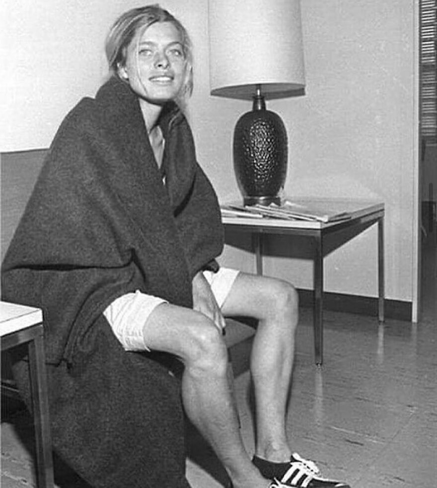 A smiling woman wrapped in a blanket, wearing shorts and sneakers, sits on a couch. Fascinating photos about history.