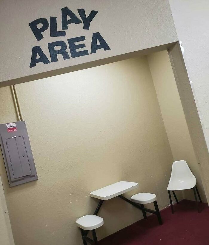 A dimly lit "Play Area" sign above a small, sad room with a table, two stools, and a single chair. A cursed image.