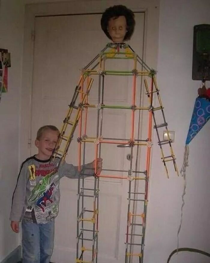 A smiling boy stands next to a life-sized stick figure with a creepy doll head, a cursed image.