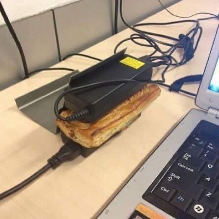 A laptop charger rests on a pastry on a wooden desk. This cursed image will confuse and amuse viewers.
