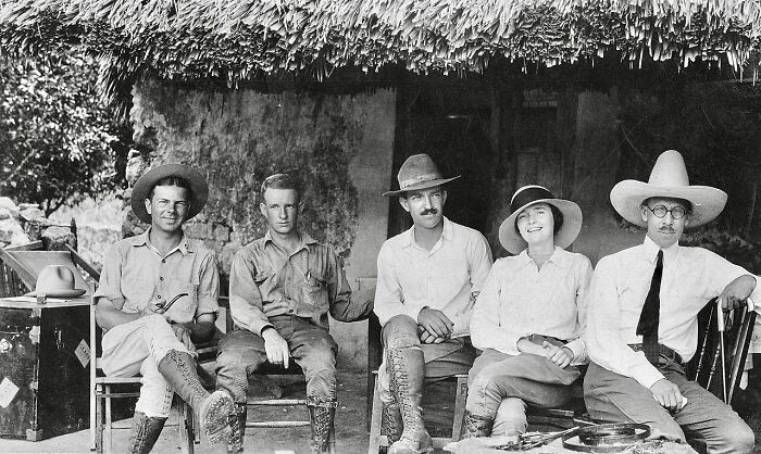 Group of people in vintage attire sitting outside a thatched-roof building in a rare and interesting historical photo.