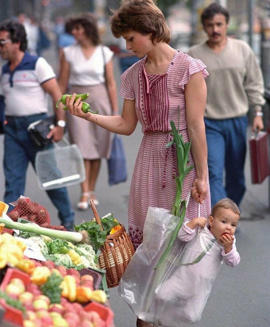 A woman in a pink striped dress holds green peppers while carrying a baby in a plastic bag with vegetables. Fascinating photos of history.