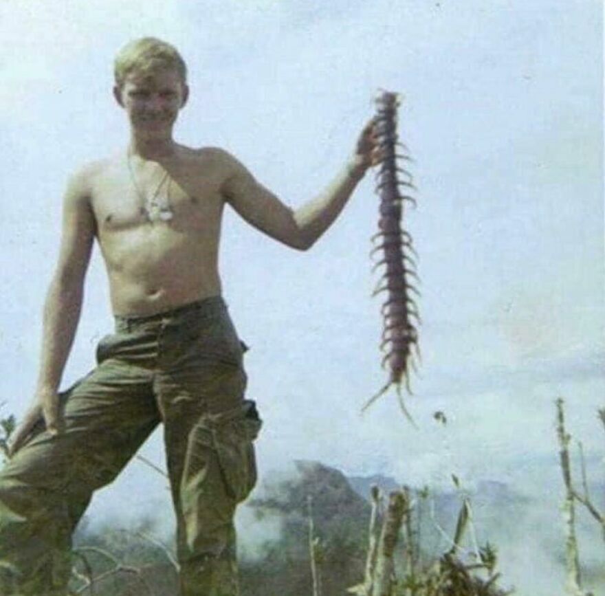 A shirtless soldier in olive green pants proudly holds up a giant centipede, offering a fascinating photo of history.