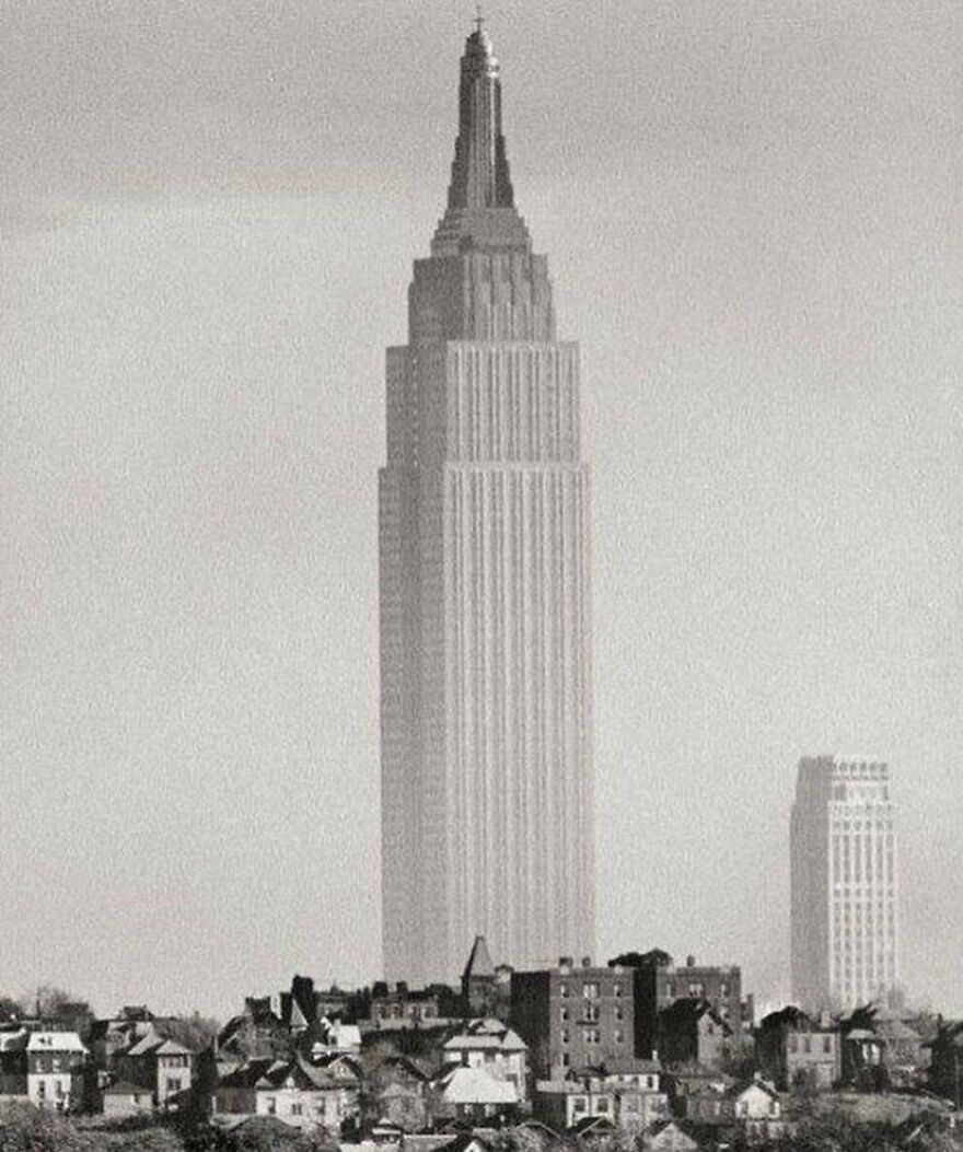 Fascinating photo of the Empire State Building towering over houses and another skyscraper, teaching about history.