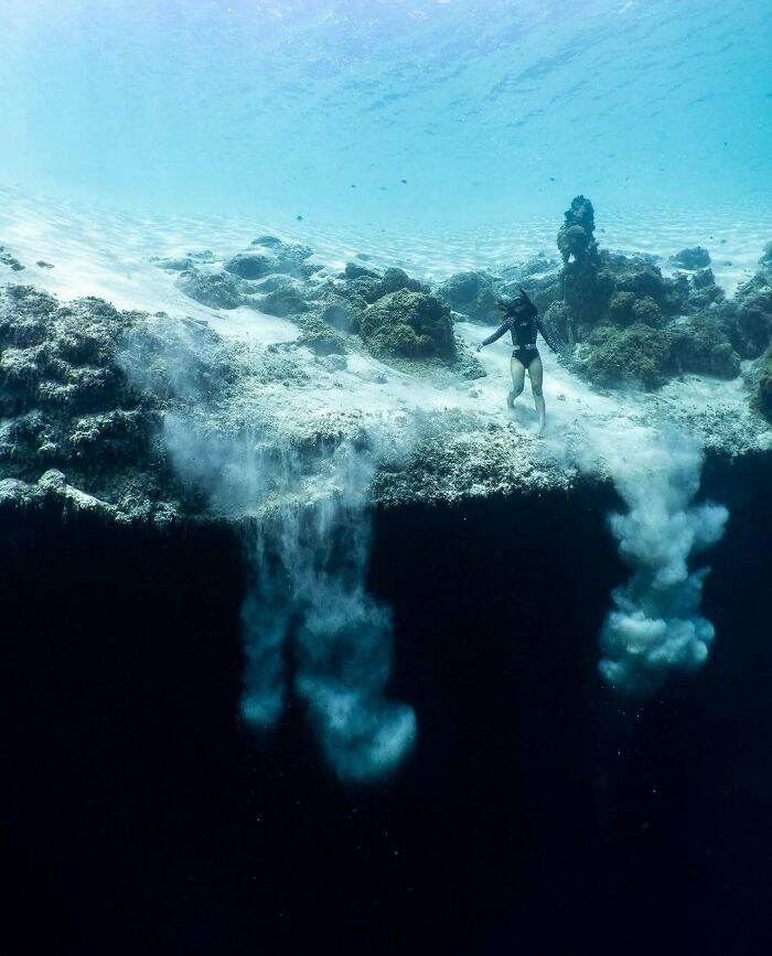 An uncomfortable water pic of a diver standing on a shallow seabed with sand falling into a deep, dark abyss.