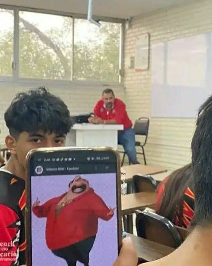 A student holds up a phone displaying an animated character wearing a red shirt, matching the teacher in the background. Unhinged pics.