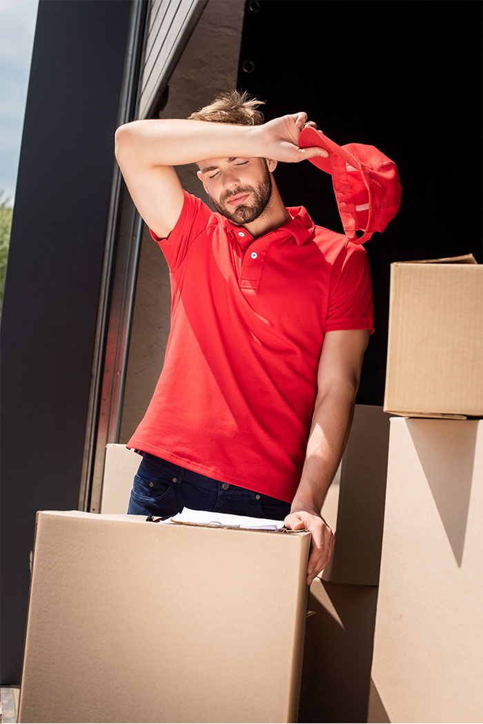 Tired delivery worker in red shirt removing cap, surrounded by boxes, illustrating overworked and underpaid friend support.
