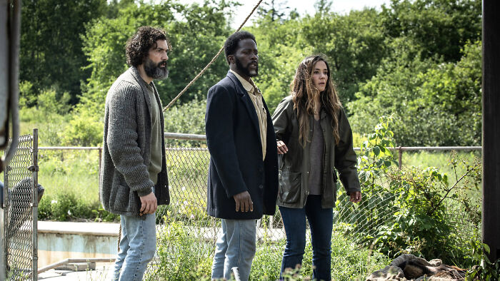 Three actors from From standing outdoors near a chain-link fence surrounded by greenery, scene from the series ending discussion.