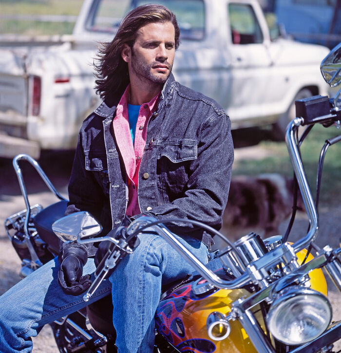 Man with long hair in denim jacket sitting on a motorcycle, evoking the style of two 80s icons now in their 60s. Man with long hair in denim jacket sitting on a motorcycle, evoking the style of two 80s icons now in their 60s.