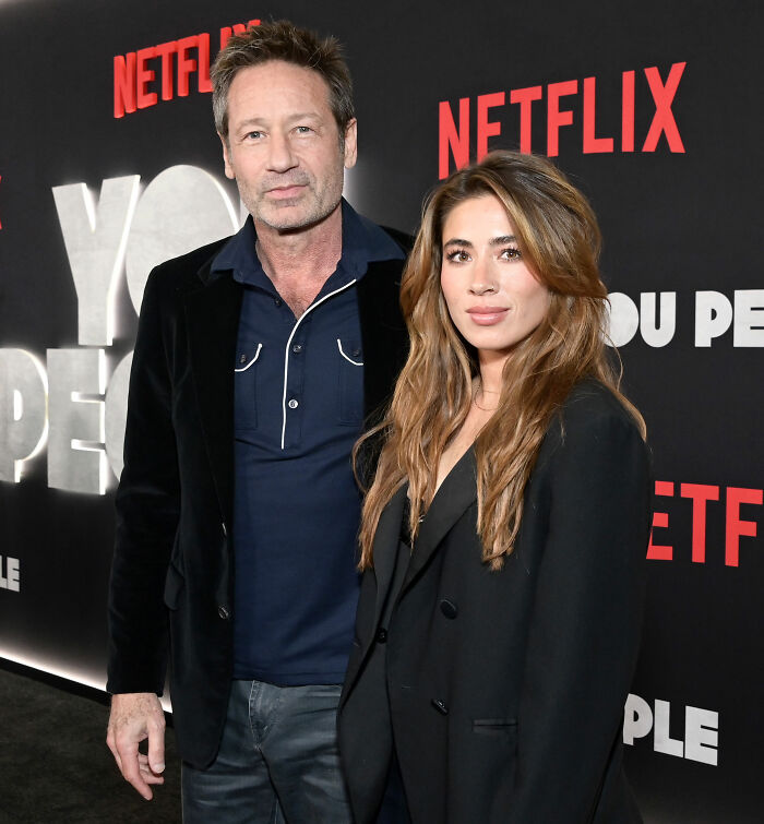 Estrela de TV dos anos 90 em um evento da Netflix posando com uma mulher, ambas vestindo roupas escuras em um cenário de marca.