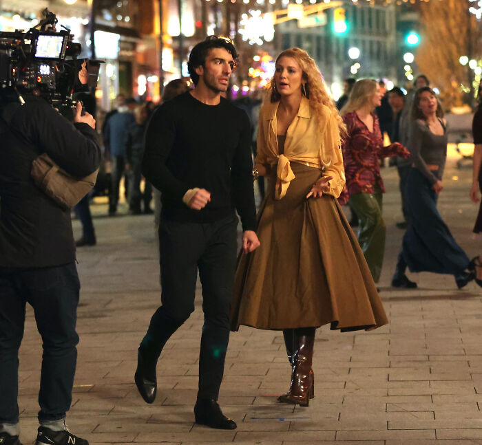 Blake Lively e Justin Baldoni filmando uma cena ao ar livre à noite, em meio às luzes da cidade e à atividade de pedestres.