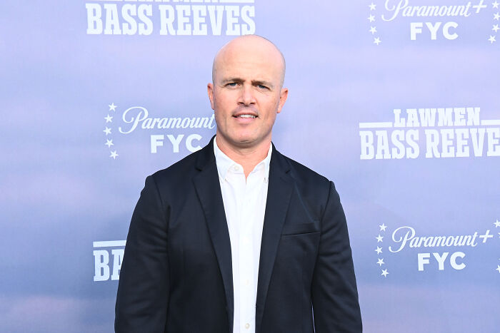 A bald man in a white shirt and black suit jacket, smiling at an event with Paramount+ FYC signage. Dutton Ranch drama related.