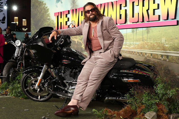 Man in a brown suit and sunglasses leans on a black motorcycle at an event, unrelated to Supergirl star Milly Alcock.