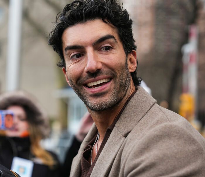 Justin Baldoni smiling outdoors, wearing a brown coat, with blurred people and city background. React explosively news.