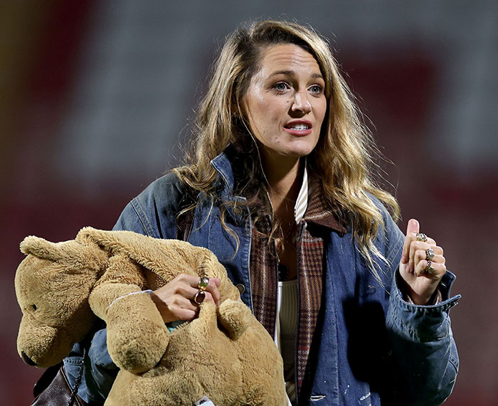 Blake Lively holding a large teddy bear, wearing a layered jacket, reacting during a public event with intense expression.