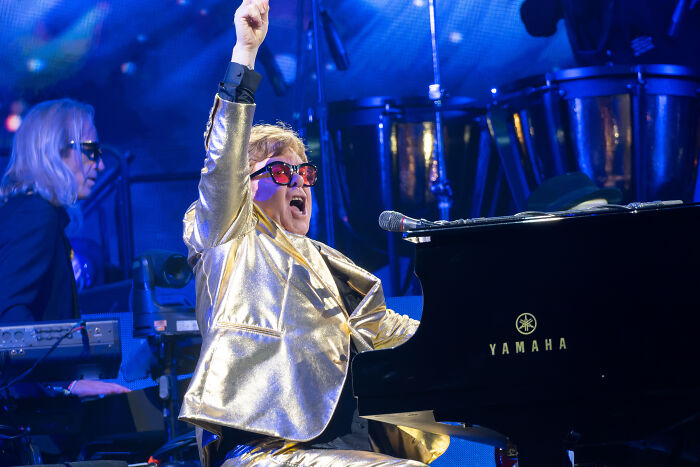 Elton John in a gold suit and red sunglasses, arm raised, playing piano. A wild rumour about this star?
