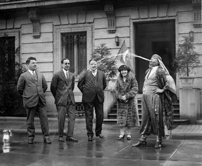 Rare and interesting photo of five people in vintage attire, one performing a fire-spitting act on a city street in the past.