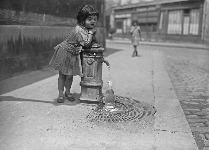 Young girl filling a bottle at a street water pump in a rare and interesting photo from a bygone era.