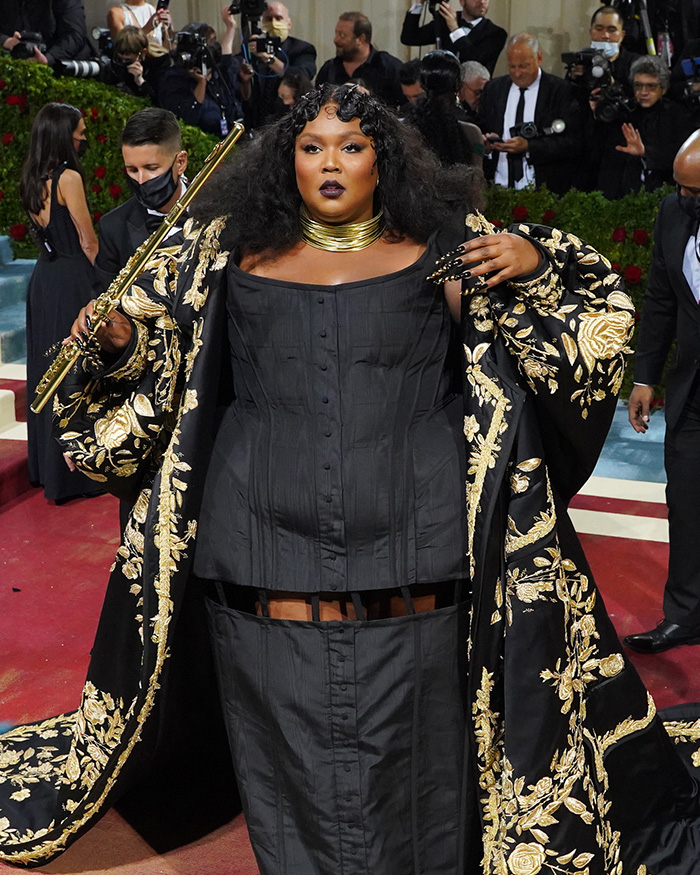 Lizzo at the Met Gala, wearing a black corset gown and embroidered robe, holding a golden flute. Celebrities often feel this way.