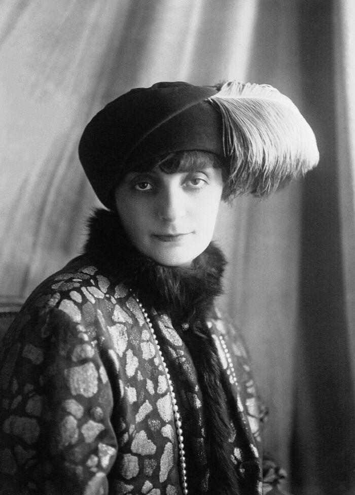 Black and white vintage portrait of a woman wearing a feathered hat and patterned coat, rare and interesting photo.