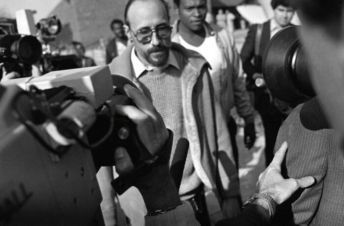 Black and white photo of a man with glasses surrounded by people and filming equipment capturing mysterious moments.