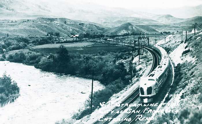 Vintage black and white image of a mysterious train traveling through a mountainous landscape at a river bend.