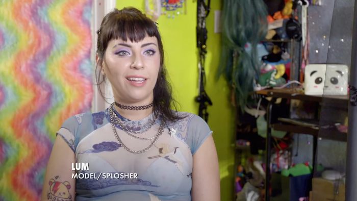 Woman with tattoos and choker sharing her experience and enjoyment of sploshing in a colorful, personal bedroom setting.