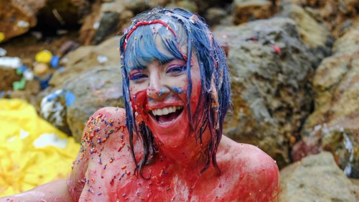 A woman covered in colorful substances, smiling and enjoying sploshing in an outdoor setting near rocks.