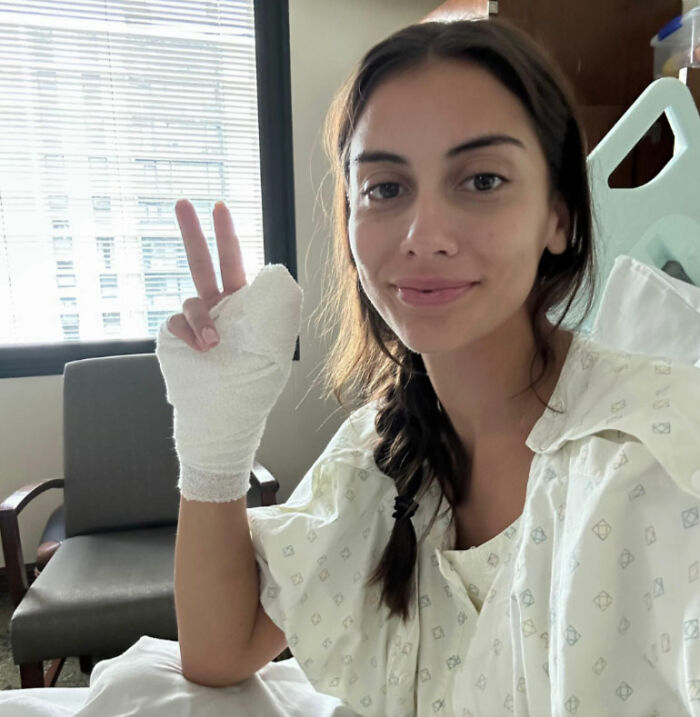 Young woman in hospital gown with bandaged hand giving peace sign, warning about risks of going to nail salons.