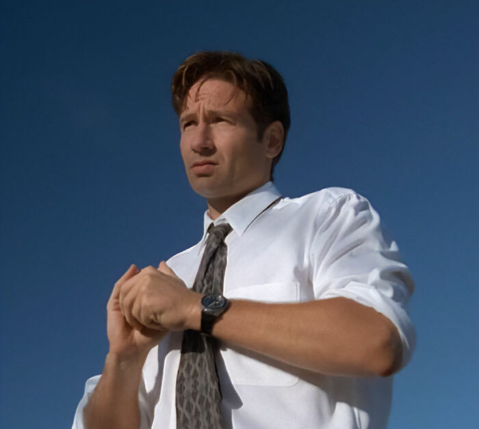 Homem de camisa branca e gravata estampada parecendo pensativo contra um céu azul claro durante um momento casual ao ar livre