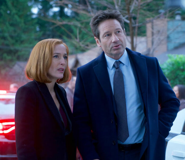 Dois atores conversando ao ar livre durante as filmagens, apresentando uma estrela de TV dos anos 90 com peruca e saia loiras.