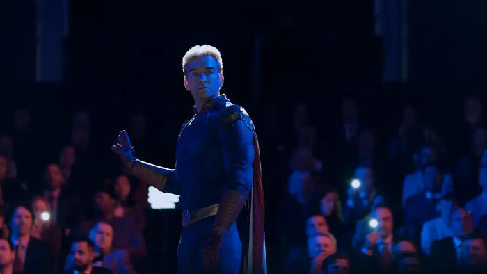 Character Homelander in a blue suit with red cape stands on stage facing an audience in a dark setting for The Boys Season 5. Character Homelander in a blue suit with red cape stands on stage facing an audience in a dark setting for The Boys Season 5.