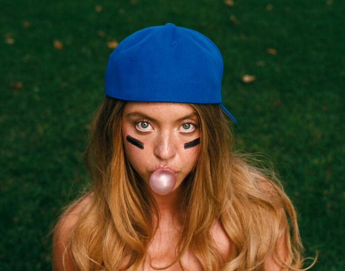 Young woman wearing a blue cap blowing a bubble gum bubble with face paint, symbolizing sploshing enthusiasm in the bedroom.
